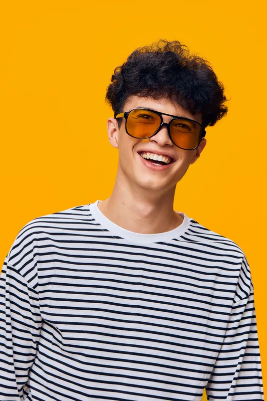 young man smiling confidently wearing yellow sunglasses and striped shirt against bright orange background, casual style with curly hair and joyful expression