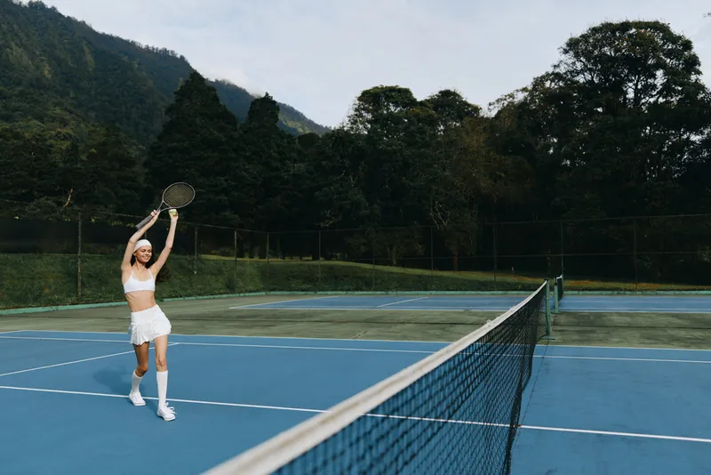 Young female tennis player in stylish white sportswear celebrates her victory on a blue tennis court surrounded by lush greenery, embodying a vibrant and energetic sports concept
