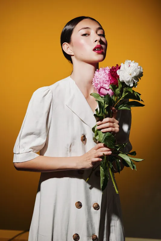 Elegant woman with red lips in light dress holding pink and white flowers. Studio portrait with warm yellow background. Concept for creative beauty and fashion.
