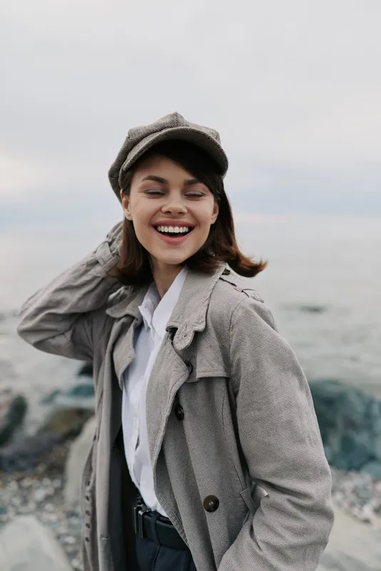 Smiling young woman in checkered coat and cap stands near rocky shore with closed eyes, enjoying fresh air and natural outdoor atmosphere by the sea on cloudy day.