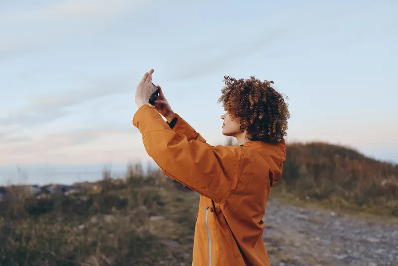 Woman lifestyle outdoors curly hair jacket moment by coast, woman smiles, arms raised to the sky at sunset, gentle breeze, relaxed posture, natural colors, scenic landscape, peaceful