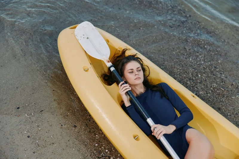 A young woman lies in a bright yellow kayak along a pebbled shore, gripping a paddle across her chest, wearing a wetsuit, under calm coastal light and tranquil water reflections.