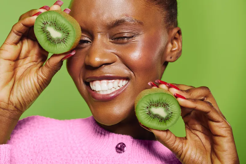Happy African woman with dark skin holds two halves of fresh kiwi fruit near her face. Smiling girl in pink sweater poses on green background. Concept of healthy lifestyle and natural food.