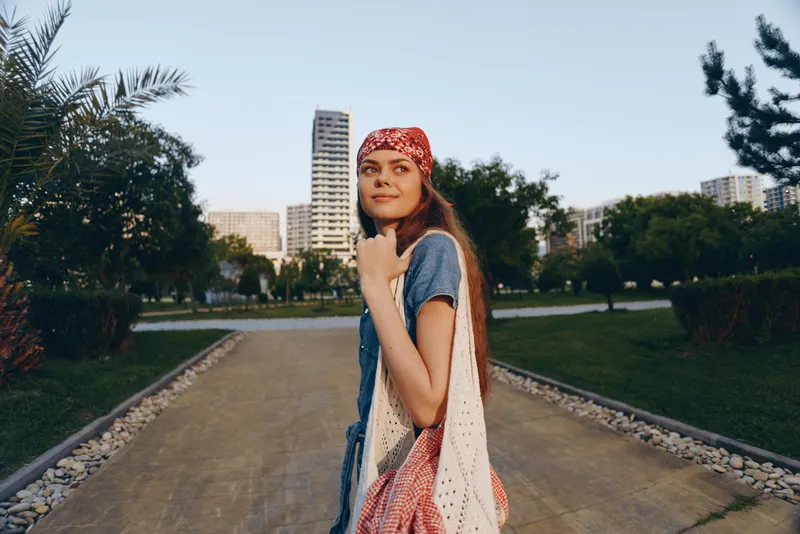 young woman with headscarf walking outdoors in summer park with greenery and urban buildings, casual style, relaxed mood, natural light