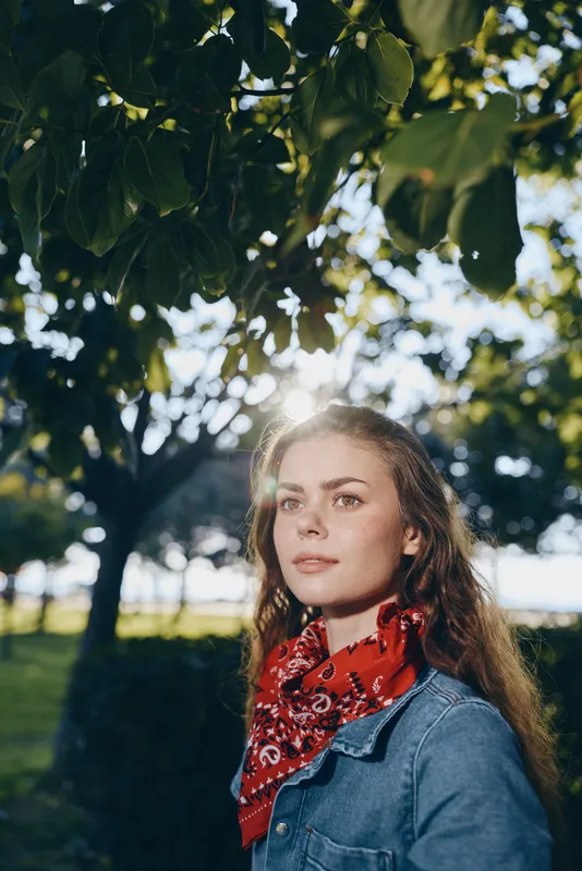 lifestyle woman wearing modern boho-western denim and red bandana in outdoor street style with warm film color sunlight and green foliage background
