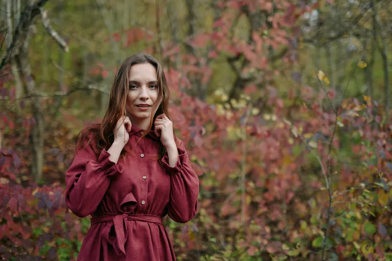 Portrait of a woman in a burgundy coat standing in a quiet autumn forest, authentic mood, natural light, candid gaze, calm posture, subtle texture, enduring style.