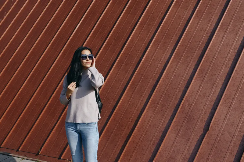 Sunglasses clad woman stands outdoors beside a slanted metal wall, casual fashion and urban attitude captured in a candid street style moment, relaxed pose, confident mood, modern backdrop.