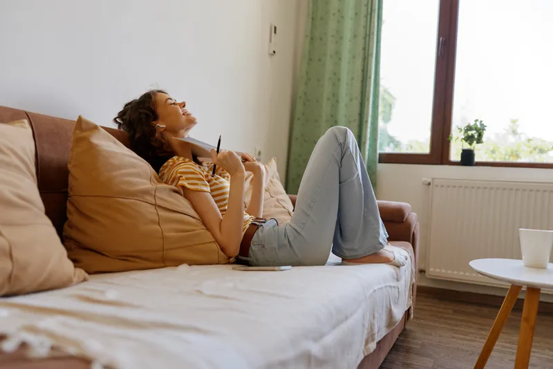 Young woman lounging on a soft sofa at home, smiling softly while resting back against cushions and clutching a warm mug, enjoying a peaceful cozy moment in a bright living room