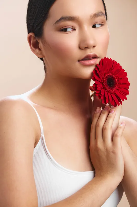 asian woman holds a vivid red flower near her face in a clean studio setting, creating a soft, elegant portrait with calm expression and gentle lighting.