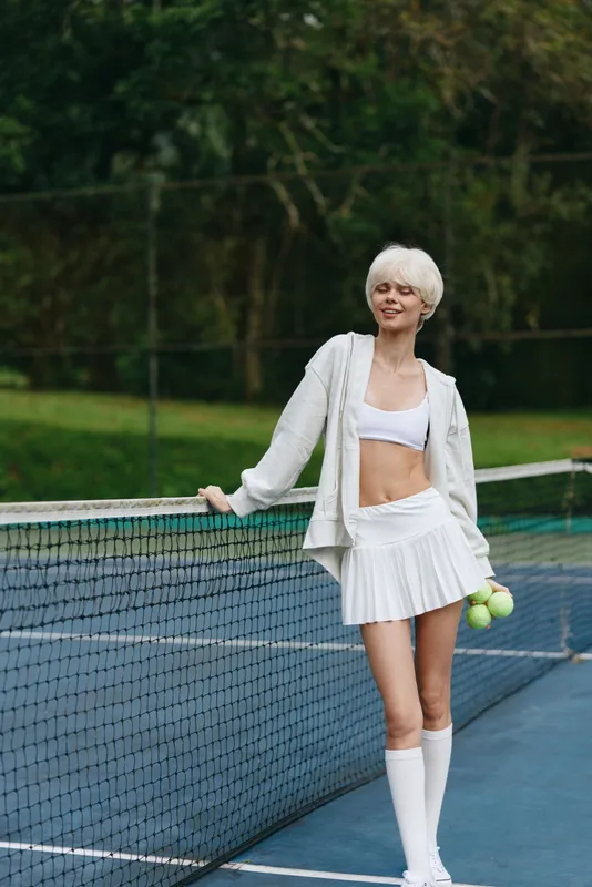 Athletic woman with short white hair in sportswear, holding tennis balls on a tennis court, exuding confidence and joy in a vibrant green outdoor setting