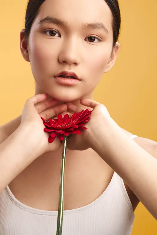 asian woman gazes softly at the camera, holding a red flower near her chin, against a warm yellow backdrop, creating an elegant, minimalist portrait.