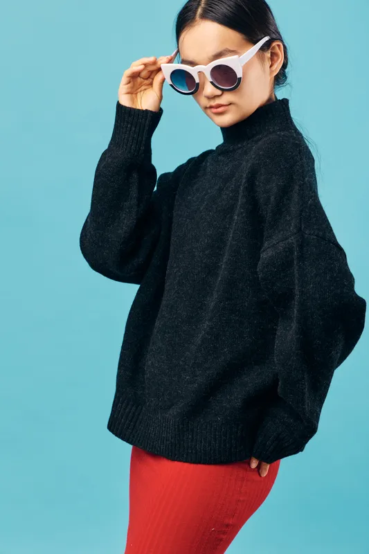Woman wearing black knitted sweater and red pants poses with white sunglasses on turquoise background. Fashion style, casual clothing, eyewear, trend and confident young female model.
