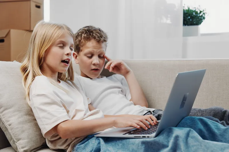 Two children sitting on a couch using a laptop together in a cozy living room with natural light. Concept of technology, learning, childhood, and family bonding at home.