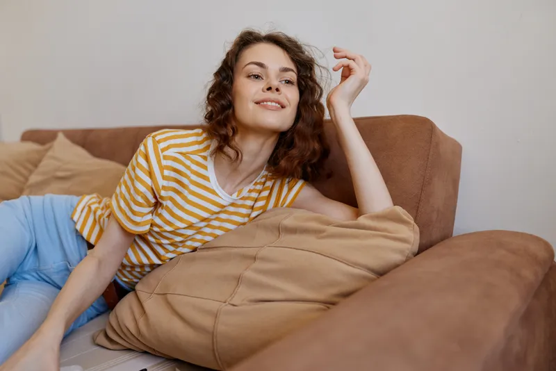 A relaxed woman lounges on a sofa in a sunny living room, wearing a yellow striped tee and jeans. Cozy home scene conveying leisure, comfort, and casual living.