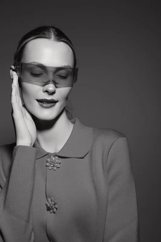 White woman with fair skin wearing translucent oversized sunglasses and vintage textured jacket poses with eyes closed and hand near face in studio. Concept of fashion and elegance.