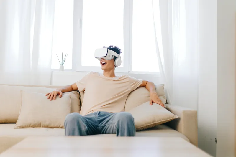 Молодой человек наслаждается опытом виртуальной реальности в шлеме VR и наушниках, удобно сидя на светлом диване в уютной, залитой солнцем гостиной, украшенной минималистичным декором