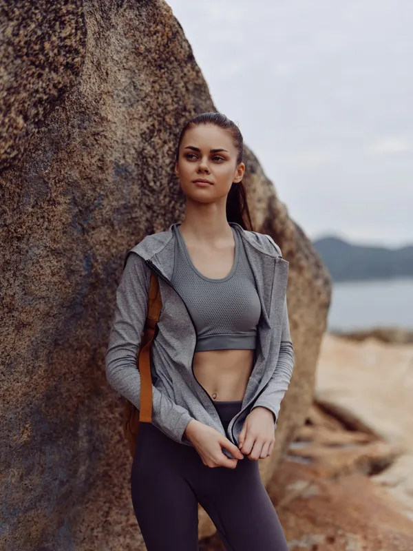 Young athletic woman in sporty attire stands confidently near a rocky shore, showcasing a healthy lifestyle and determination with a serene natural background