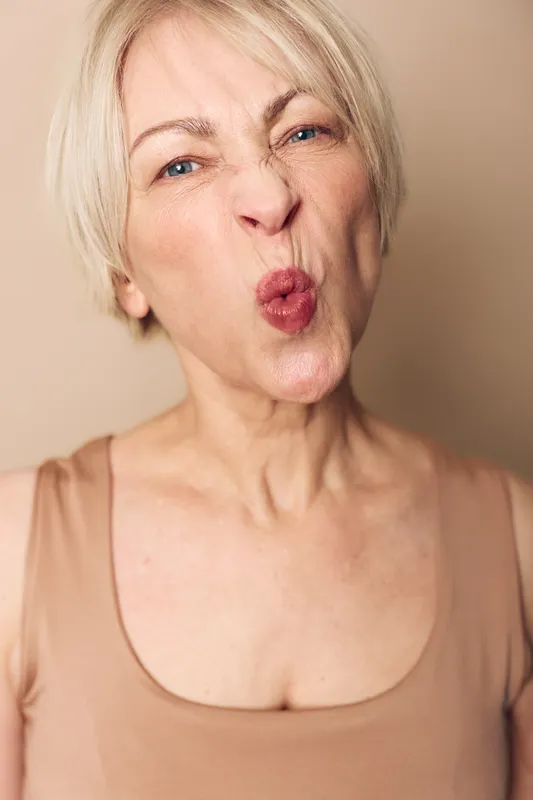Close-up of an elderly woman making a playful face with puckered lips and squinted eyes. Natural skin texture and short blond hair visible against neutral background.