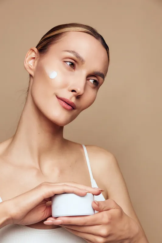 A young woman with clear skin applies moisturizing cream on her cheek while holding a white jar, posing against a beige background. Concept for skincare, beauty, and self-care routines.