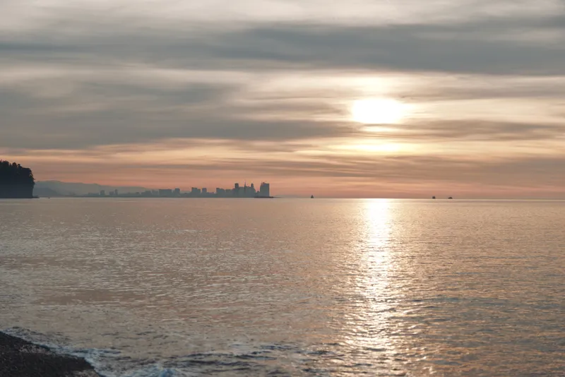 Sunset over calm water with a distant city skyline on the horizon, gentle reflections on the sea, and a quiet shore framing a serene evening seascape.