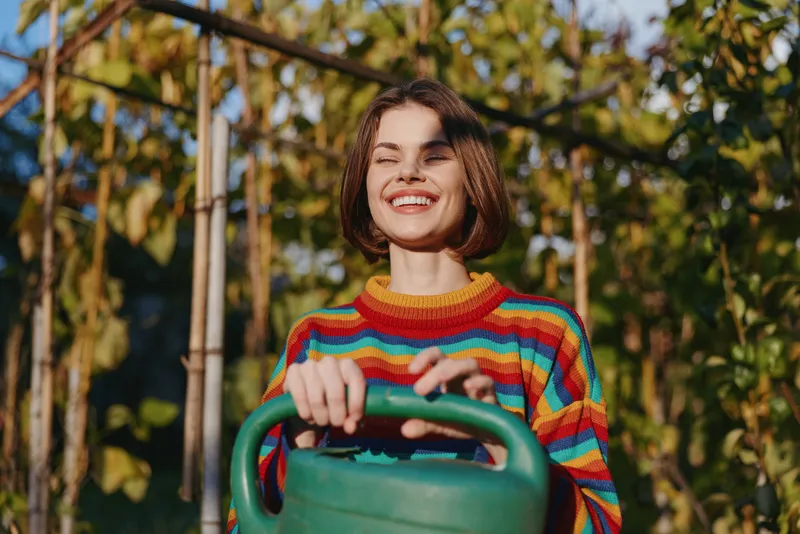 Woman in a colorful striped sweater smiling while holding a green watering can in a sunlit garden by a wooden trellis, short bob hair and joyful outdoor lifestyle scene.