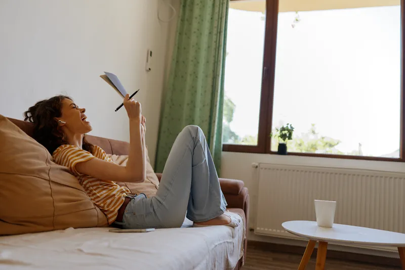 Young woman relaxes at home on a couch, writing in a notebook while smiling, enjoying a casual lifestyle moment in a sunlit living room.