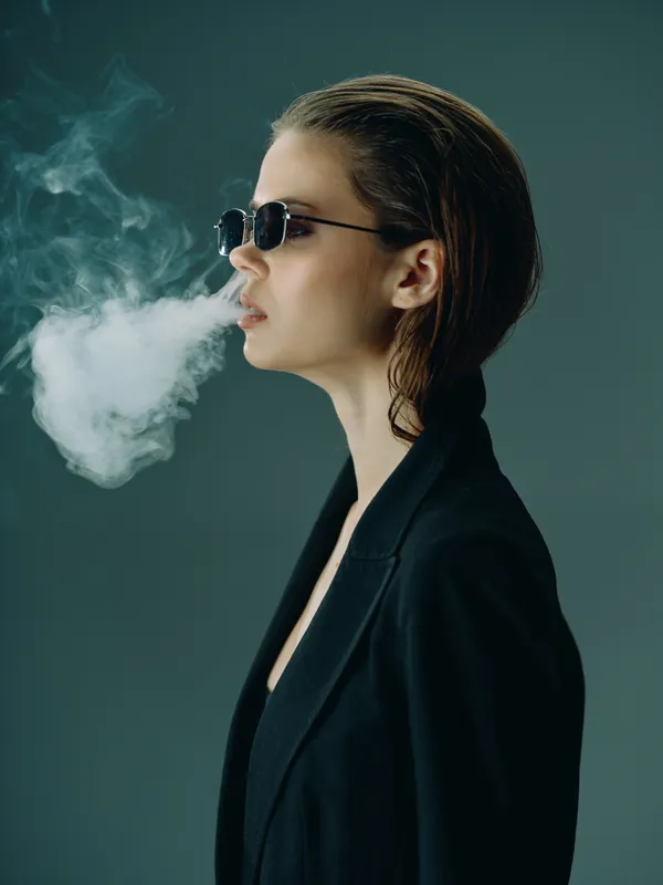 Young woman in sunglasses smoking with visible smoke cloud on dark background, stylish and cool concept.