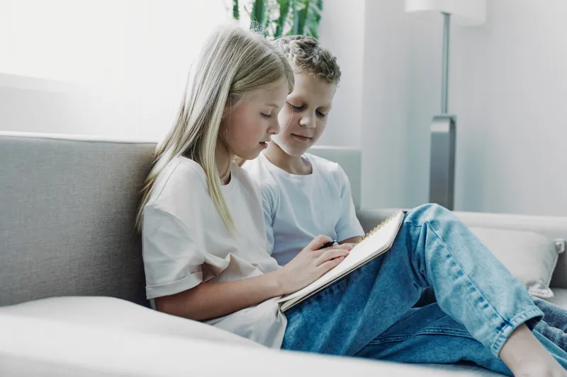 Two children sitting on a sofa, focused on drawing in a notebook. Casual clothing, natural light, cozy home environment. Concept of creativity, learning, and childhood bonding.