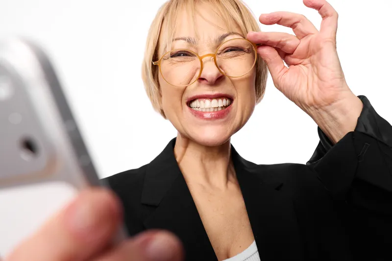 Woman business glasses smile adjusting phone selfie senior indoor closeup, older woman laughing while adjusting eyeglasses, holding smartphone, clean white background, professional mood
