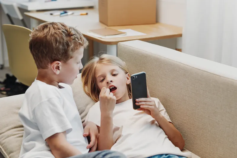 Two children relax on a beige sofa in casual clothes, one holding a smartphone and showing something to the other. Cozy home environment.