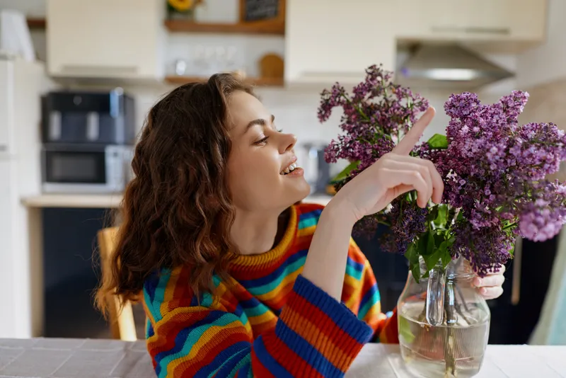 Woman in a colorful striped sweater arranges fresh flowers in a glass vase at home, savoring a cozy indoor moment while creating a cheerful bouquet on a kitchen table.