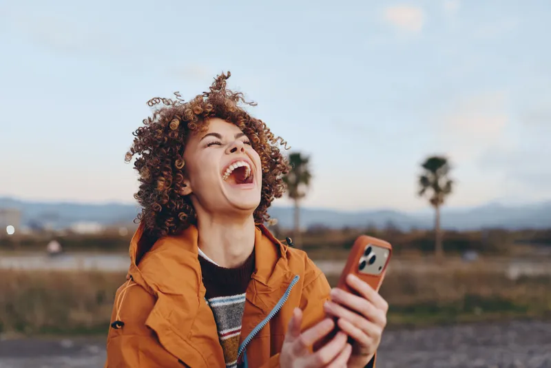 Outdoors lifestyle photo of a woman with a smile, curly hair, holding a smartphone for a selfie. She wears an orange jacket, sunset sky and soft field blur behind, in warm light today.