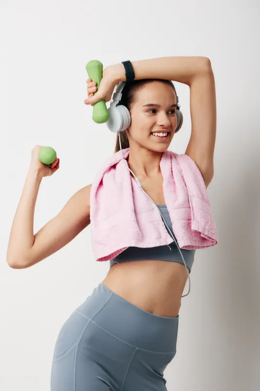 Young woman in athletic wear stretching with light green dumbbells, wearing over-ear headphones and a pink towel draped over her shoulders, smiling against a clean white studio background, fitness