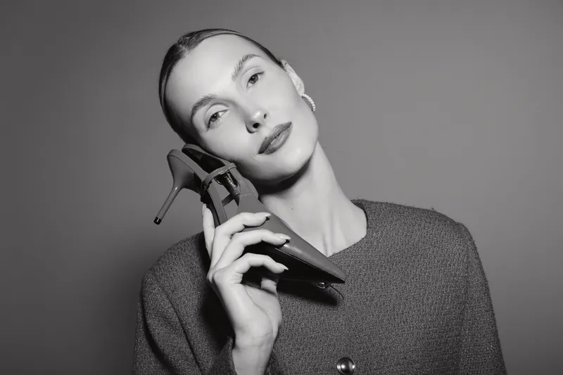 Elegant white woman with pale skin holds high heel shoe near face against grey background. Concept of fashion, footwear, and style.