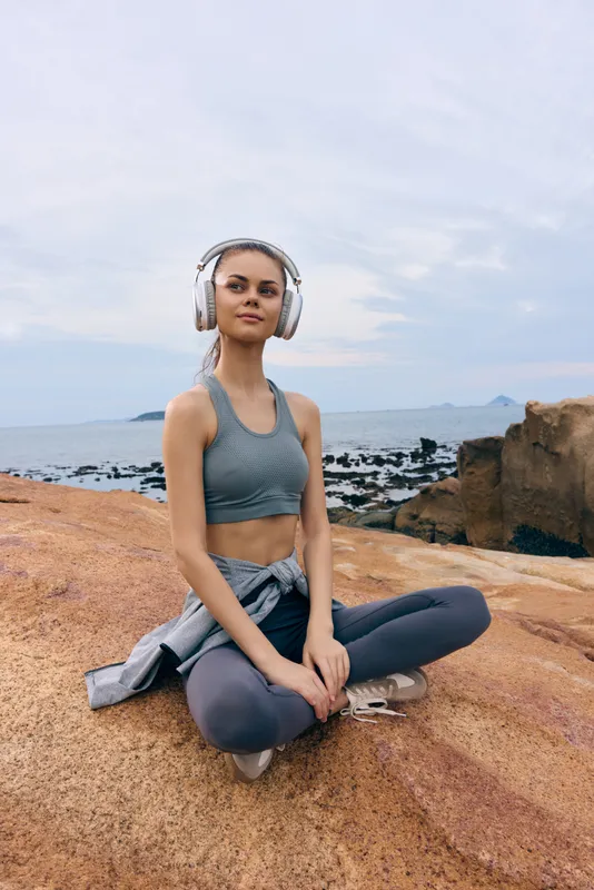 Young woman practicing mindfulness and meditation on rocky beach, wearing sportswear and headphones, embodying tranquility and health with a serene coastal background