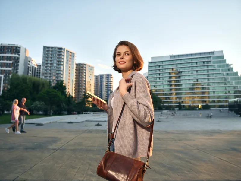 A cheerful professional woman stands outdoors in an urban setting with modern buildings in the background. She smiles warmly, carrying a brown purse, exuding confidence.