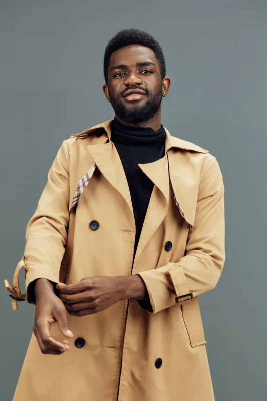 Stylish black male model wearing a beige trench coat on a gray background, showcasing confidence and modern fashion trends