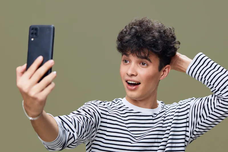 young man taking selfie with surprised expression wearing striped shirt, casual style against neutral olive background in studio lighting