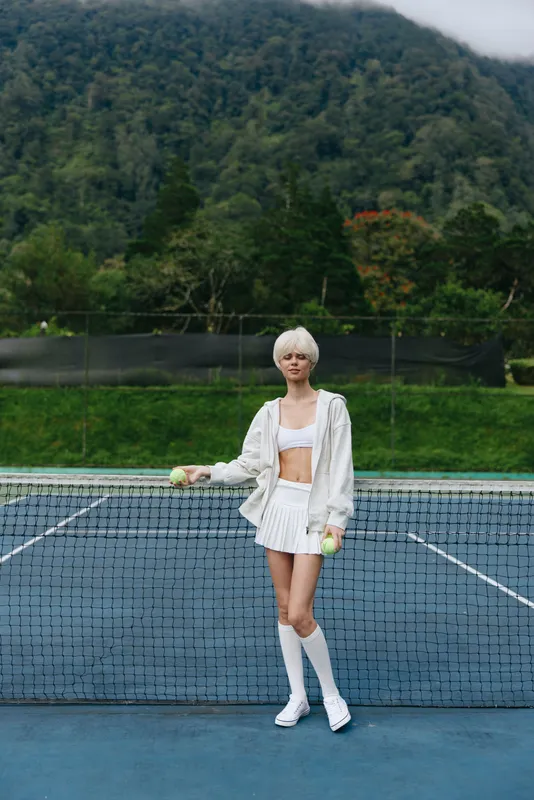 Young female athlete in white sportswear poses confidently on a tennis court, showcasing determination and focus against a lush green background