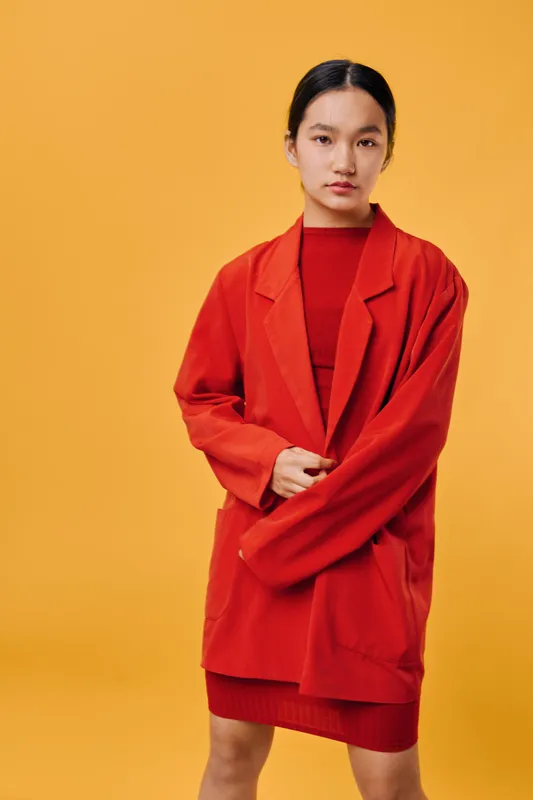 Young woman wearing red oversized blazer and dress standing with serious expression on yellow background. Fashion model with dark hair and minimal makeup posing in studio with hands crossed.