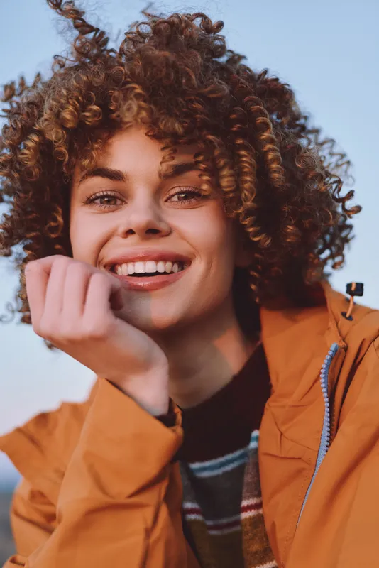 Curly hair woman smile lifestyle outdoors portrait casual jacket in warm daylight closeup view of young adult touching cheek and laughing natural beauty soft wind friendly urban street