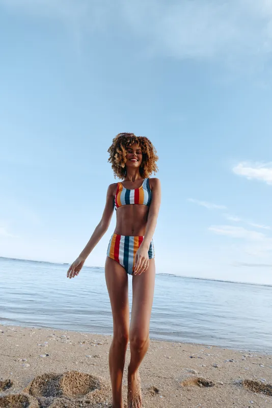 Young woman in colorful striped bikini stands on sandy beach near calm sea under blue sky, enjoying summer vacation and warm sunny weather outdoors.