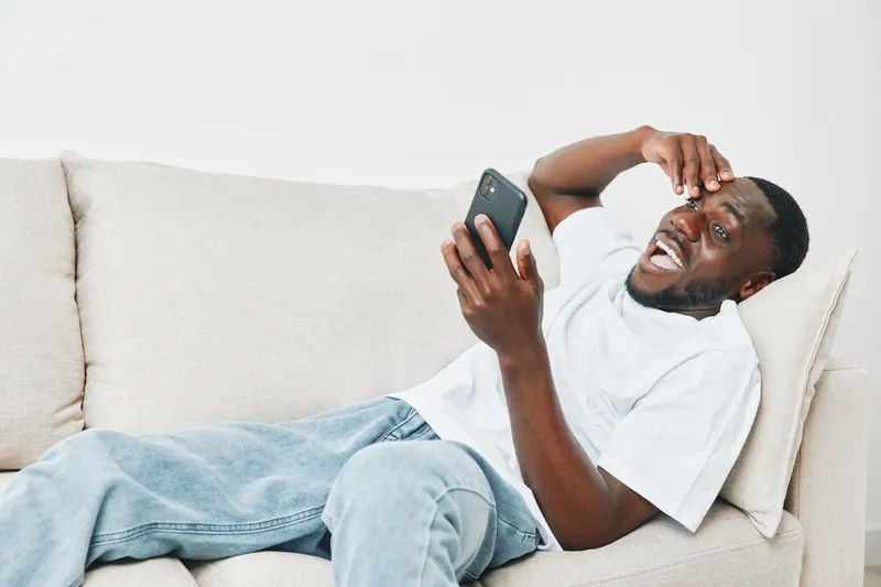 A young Black man expresses frustration while using his smartphone on a couch, showcasing emotions of surprise and confusion