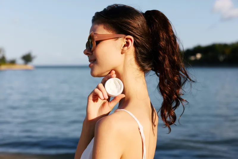 A stylish woman stands beside calm water wearing sunglasses and a white swimsuit strap, gently holding a shell at her neck in a sunlit, serene moment for a relaxed coastal mood.