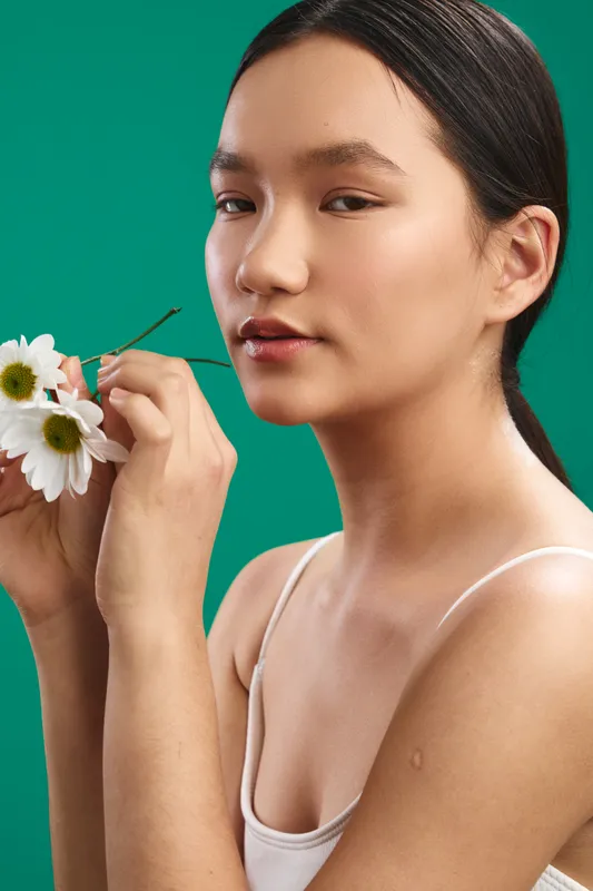 asian woman holds white flowers against a vivid green backdrop, gazing softly at the camera with a serene, confident expression that conveys natural beauty, calm mood, and fresh elegance.
