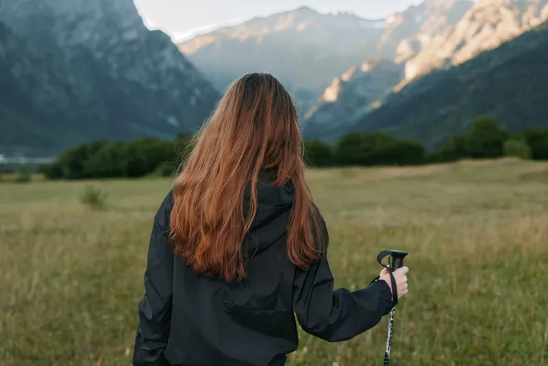 A lone hiker stands in a wide meadow facing distant mountains. She wears a dark jacket and grips a trekking pole, enjoying a peaceful outdoor moment.
