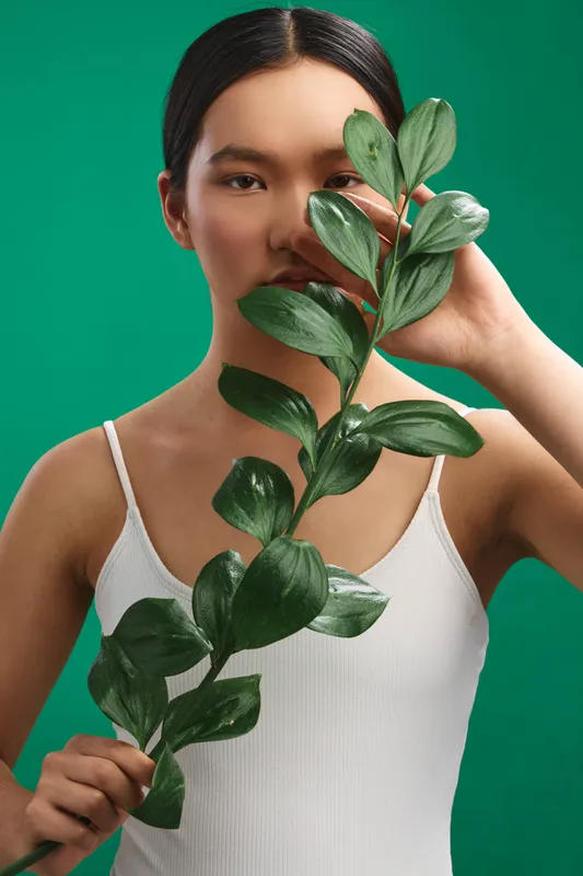 asian woman holds a lush plant with broad leaves, partially covering her face in a studio portrait against a vivid green backdrop, creating a calm, natural, contemporary mood that emphasizes balance