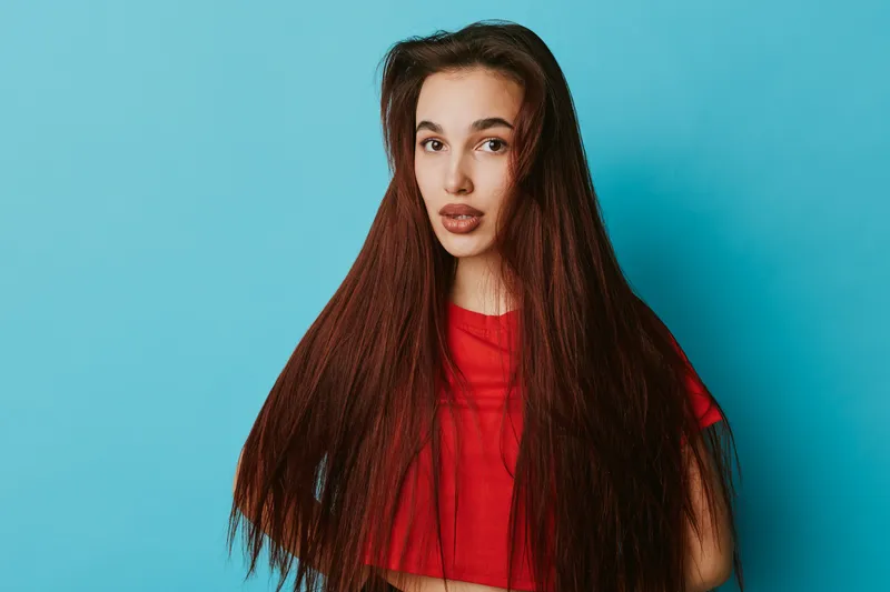 Young woman with long straight hair and red shirt posing on blue background looking at camera with natural expression for beauty, fashion and portrait concept.