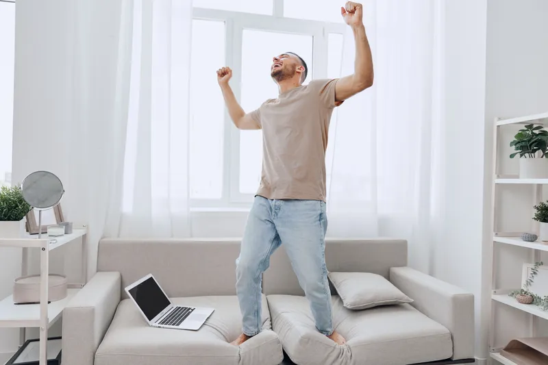 Happy man celebrating work-life balance while standing on the couch in a cozy home office environment, reflecting modern challenges and self-care.