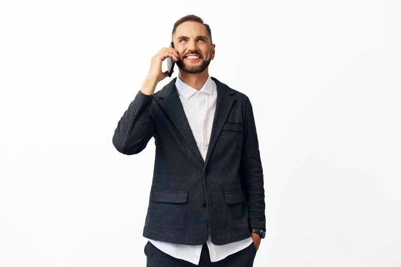 A confident business professional man stands against an isolated colored background, speaking on a phone with a friendly smile, exuding poise, focus, and readiness for discussion.